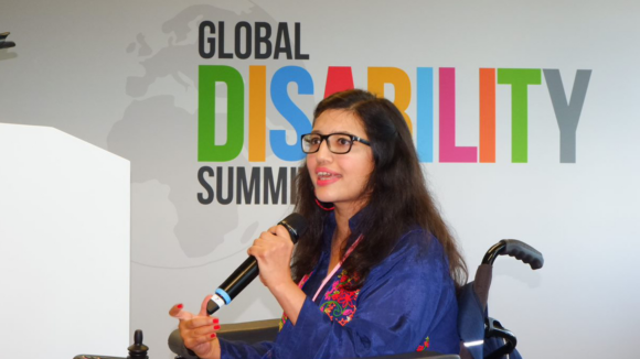 Disability activist Abia Akram speaking into a microphone from her wheelchair on stage at the Global Disability Summit