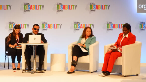 Four people sit on a stage in front of a backdrop featuring the words 'Global Disability Summit'.