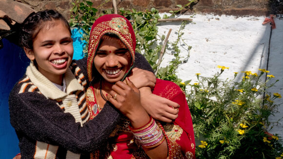 Two women smile and hug: both are wearing colourful clothes. One has a visual impairment.