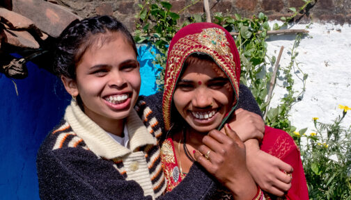 Two women smile and hug: both are wearing colourful clothes. One has a visual impairment.