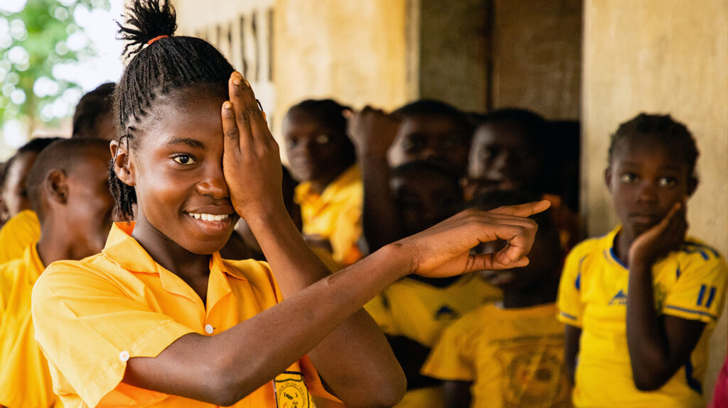 A girl smiles and points while covering her left eye with her hand.