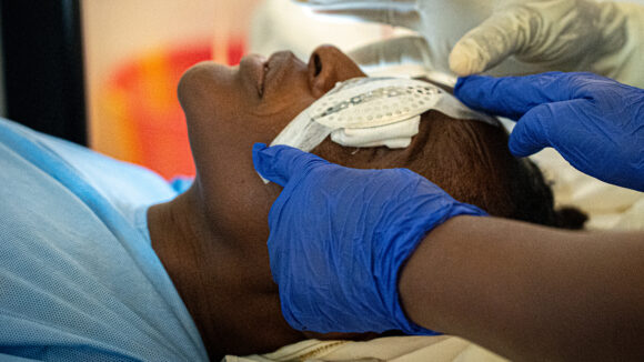 A young person is lying back on a hospital bed, having a bandage placed on their left eye.