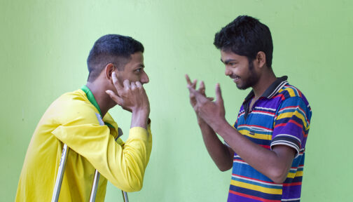 Two young men speak to each other in sign language. One wears a yellow t-shirt and holds crutches, the other wears a blue striped t-shirt.