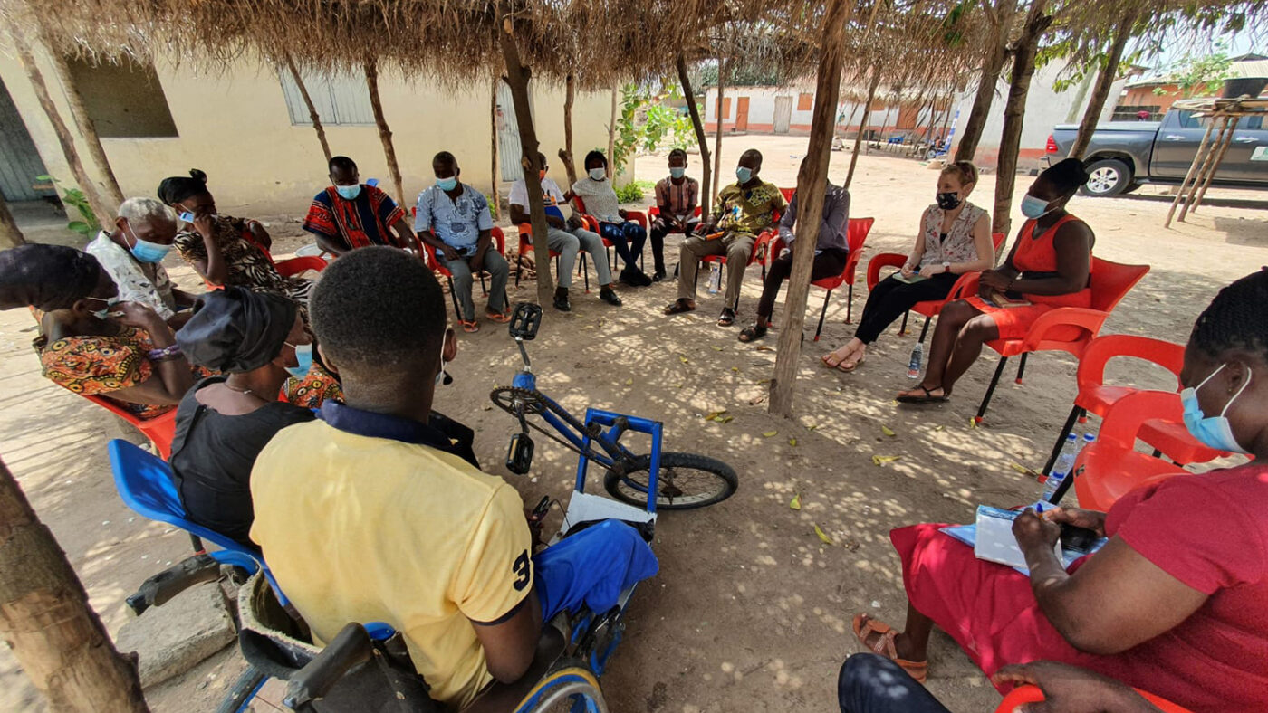 A large group of people sit in a circle outside in a shaded area next to a building.