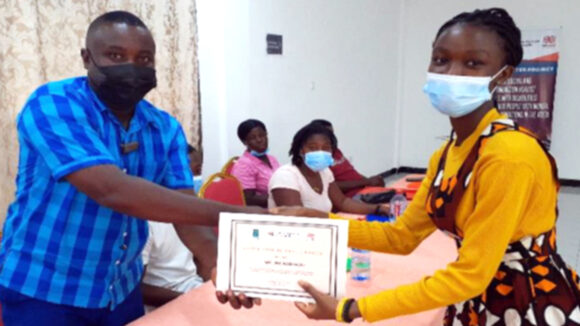 A man shakes hands and gives a woman a certificate for completing her inclusion ambassador training. Behind them at a table, three women look on.