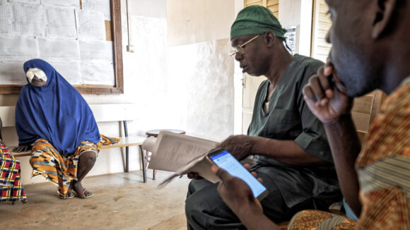 Two health workers, one wearing scrubs, look at papers and a mobile phone while gathering data. A patient with her eyes bandaged sits in the background.