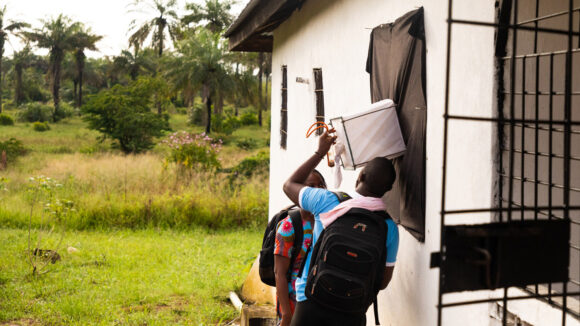 A man and a woman stand next to a house checking a mosquito trap, which is attached to the exterior wall..