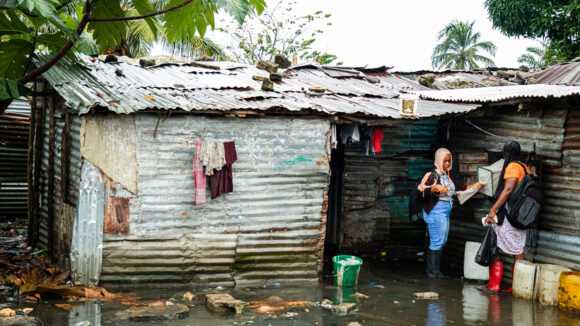 Two women stand in a flooded area outside a house, where they are checking a mosquito trap.