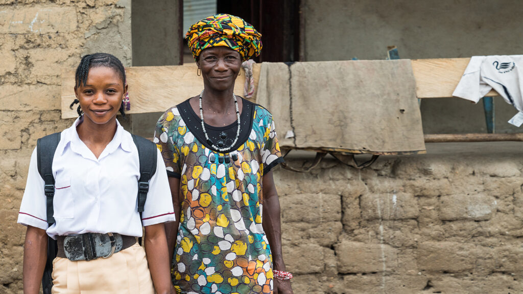 Saio and her mother stand together outside their home. They are both smiling.