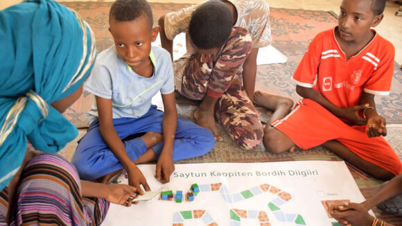 A group of children sit around a board game on the floor.