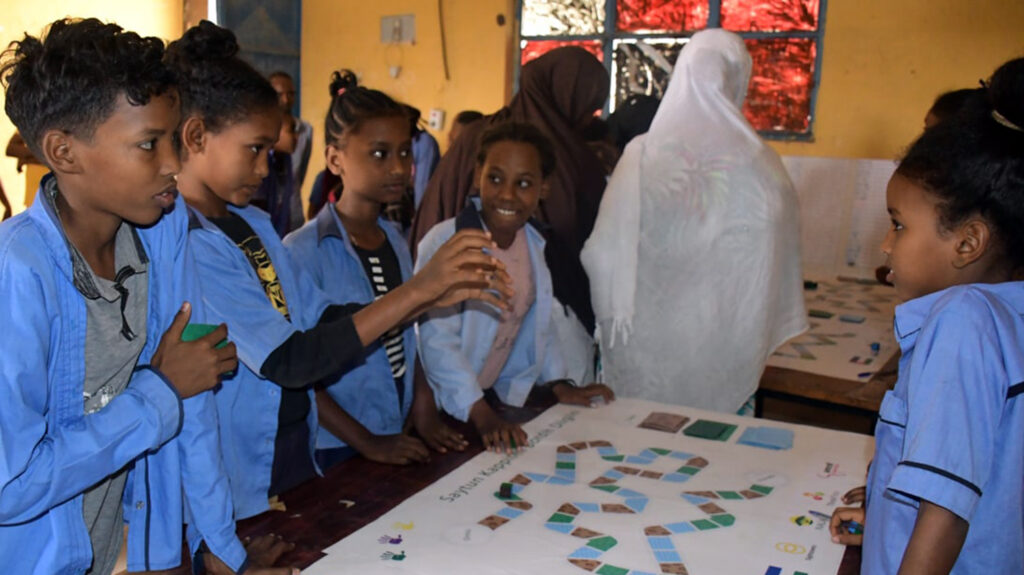 Schoolchildren stand either side of a table on which they are playing a board game.
