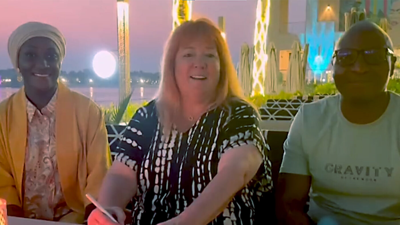 Sightsavers CEO Caroline Harper smiles alongside two colleagues. She's holding a pen. In the background are some brightly lit skyscrapers.
