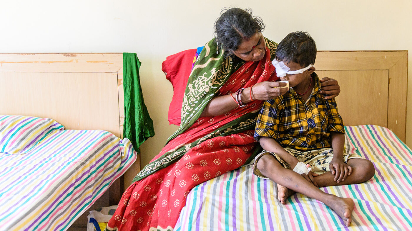 Sanjit sits with his mother on a bed in the hospital. He has a bandage over his right eye, and his mother is giving him a sip of water.