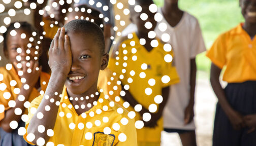 A school student wearing a bright yellow shirt smiles as he holds his right hand over his eye. There's an illustrated ring of small dots surrounding his face.