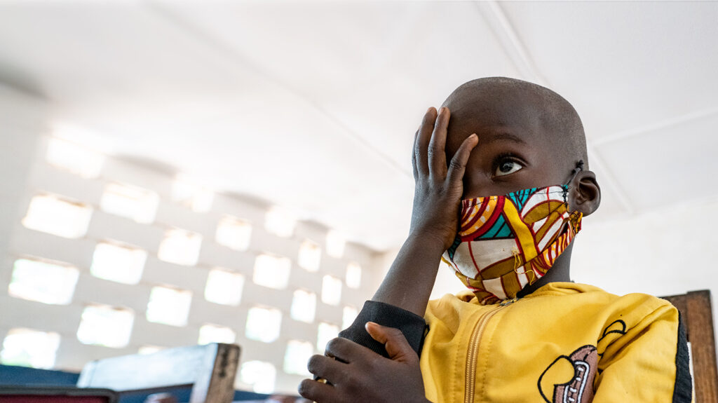 A young boy wearing a colourful face mask and a bright yellow jumper covers hish right eye with his hand during an eye test.