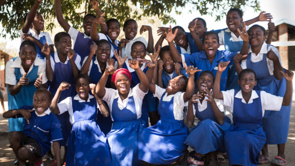 A large group of schoolchildren in uniform smiles and cheers.