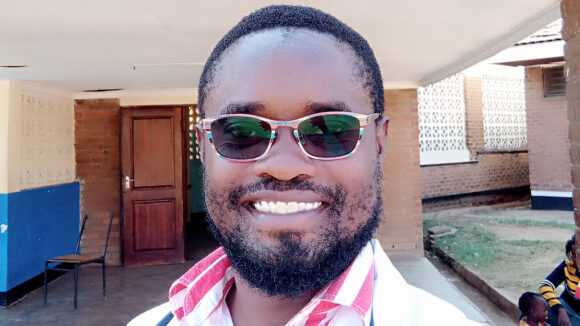Alinafe stands outside a hospital smiling while wearing sunglasses.