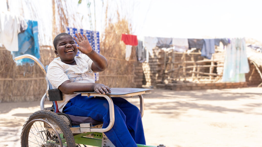 in Malawi, a young boy in a wheelchair smiles broadly and holds his hand up to wave at the camera.