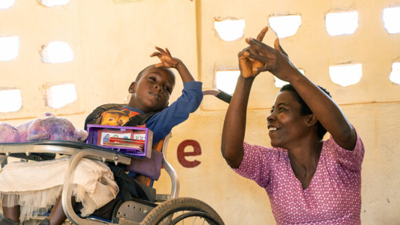 Nora in her classroom with a student who is in a wheelchair. Nora is holding her hands in the air, and the student is watching.