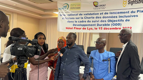 During a press interview in Senegal, a man faces a video camera while speaking into a microphone. Three fellow event attendees are standing behind him.