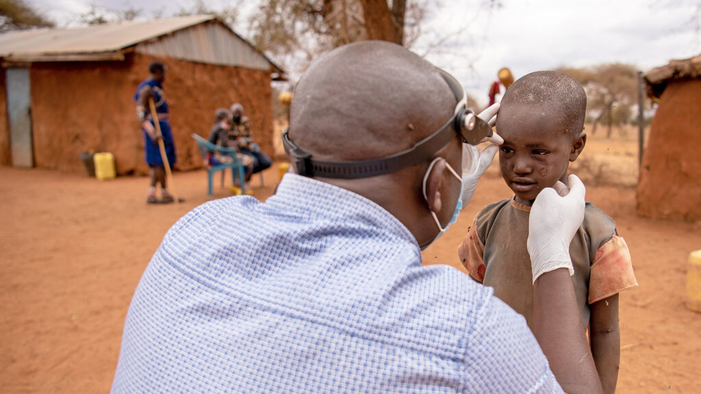 A child in Kenya has his eyes checked by an eye health worker wearing gloves and a mask. They're outside in a village with dusty red sand on the ground.