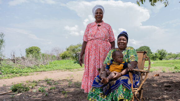 Holo sits in a chair outside, with her young grandson on her lap. Her daughter Rachel stands next to them with her hand placed on Holo's shoulder.
