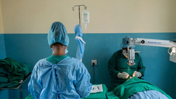 An eye surgeon operates on a patient in hospital. Another member of clinical staff stands next to them wearing scrubs.