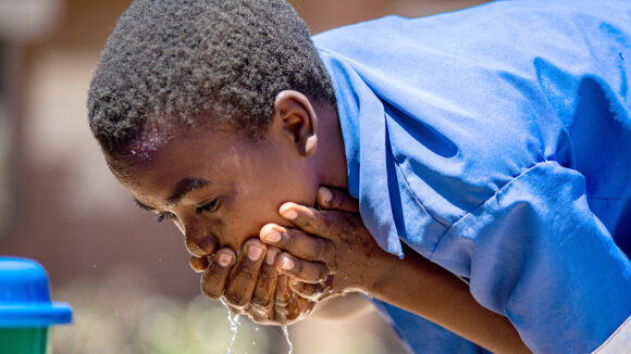 A boy wearing a bright blue shirt washes his face using water from a tap.
