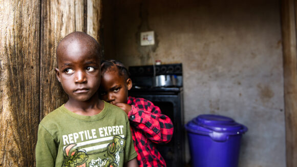 A young boy and his sibling standing in their home, looking solemn.