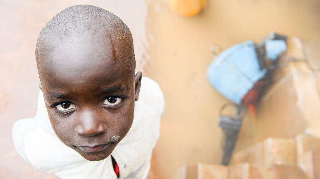 11-year-old Emmanuel looks up at the camera. He's standing on dusty ground with muddy puddles.