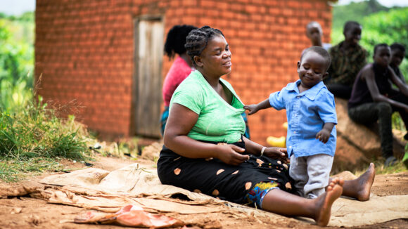 One-year-old Mobwin stands at his mother's feet as she sits outside their home in Zambia. The floor is dusty and there are brick buildings in the background.