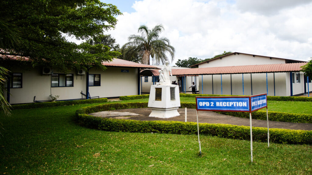 A hospital building in Zambia, featuring concrete buildings with corrugated roofs, set among lush trees, hedges and grass.