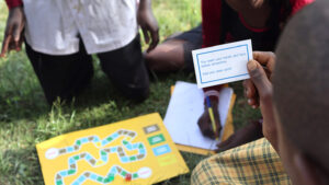 School children sitting on the grass playing a board game. One is holding up a card that says: 'You wash your hands and face before dinnertime. Add one clean point!'