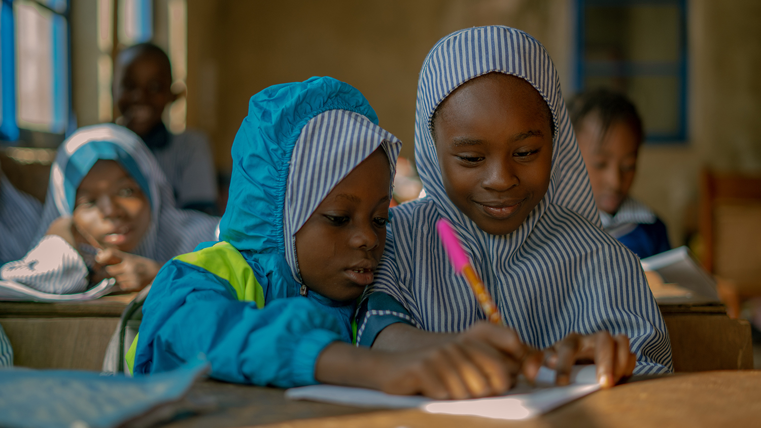 Nine-year-old Khadijah at school with her friend after successful cataract surgery. They're sitting at a desk writing on a shared piece of paper, wearing their blue school uniforms.