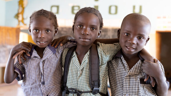 Three young school student sit with their arms round each other at school in Zambia. They're wearing checked school shirts.