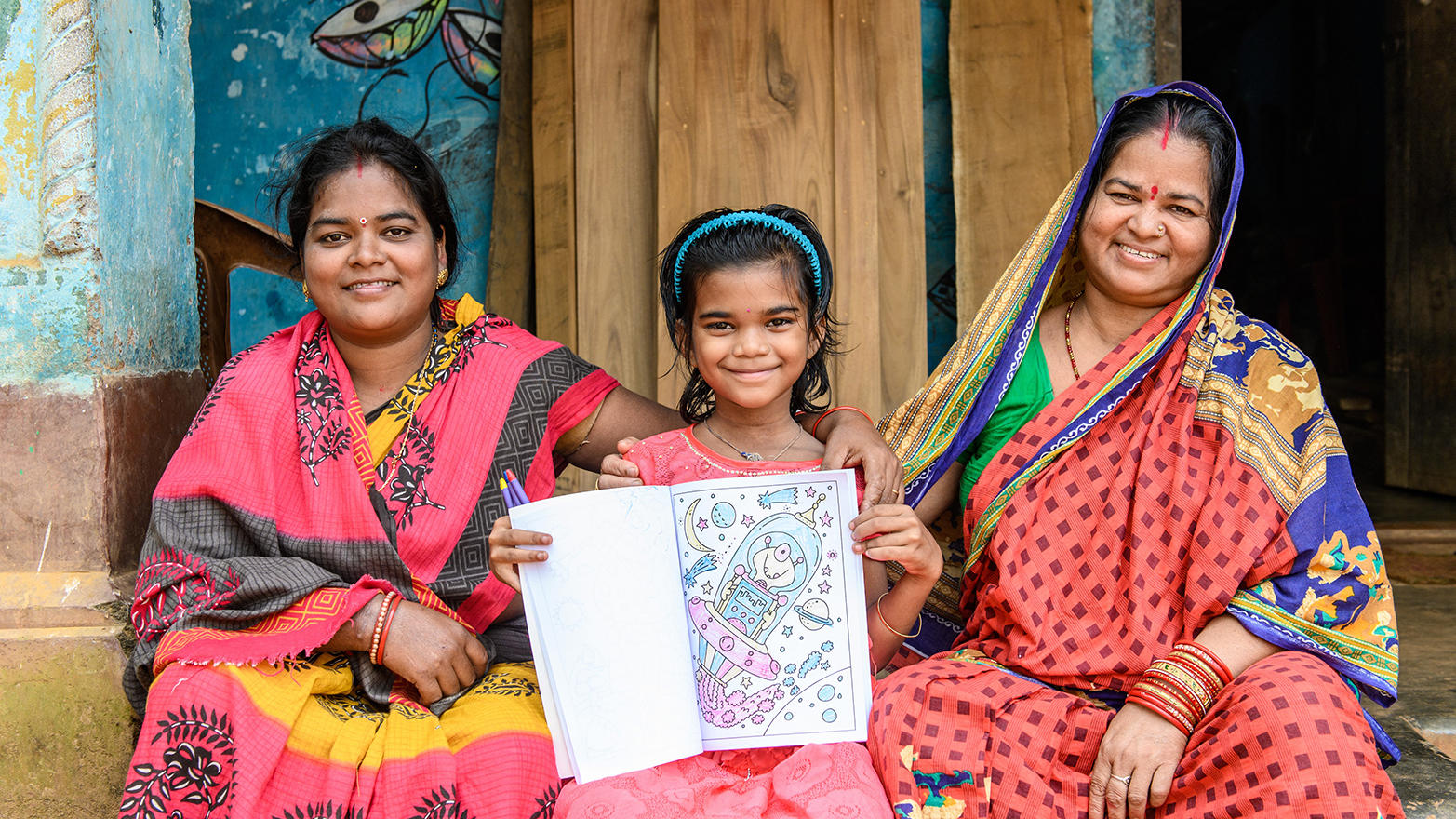 Archana sits between her mother and grandmother while holding up a colouring book.