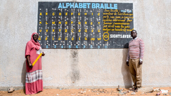 Two teachers stand either side of a colourful blackboard showing the Braille alphabet.