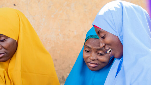 Two women wearing colourful headscarves lean together and smile.