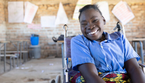 A girl smiles in a classroom in Zambia. She's sitting in a wheelchair and wearing a blue school shirt.