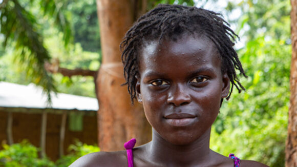 A teen girl looks into the camera standing outside under lush green trees.
