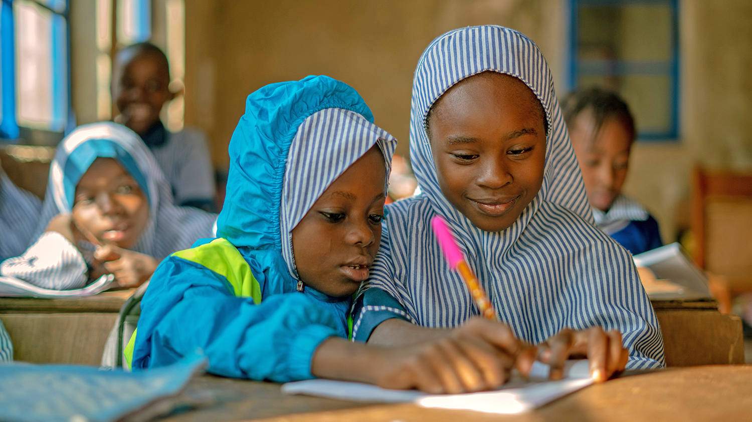 Nine-year-old Khadijah at school with her friend after successful cataract surgery. They're sitting at a desk writing on a shared piece of paper, wearing their blue school uniforms.