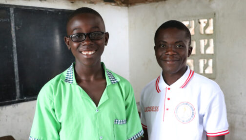 Twelve-year-old Arthur smiles as he stands next to his teacher, Mr Thompson. They stand in front of a chalk board in a classroom.