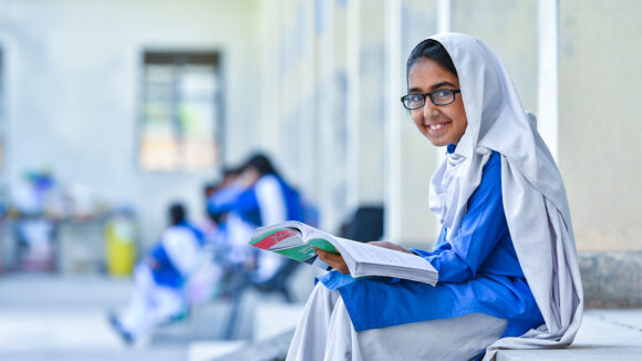 Minahil, a 12-year-old student from Pakistan, smiling while looking towards the camera and holding a school textbook.