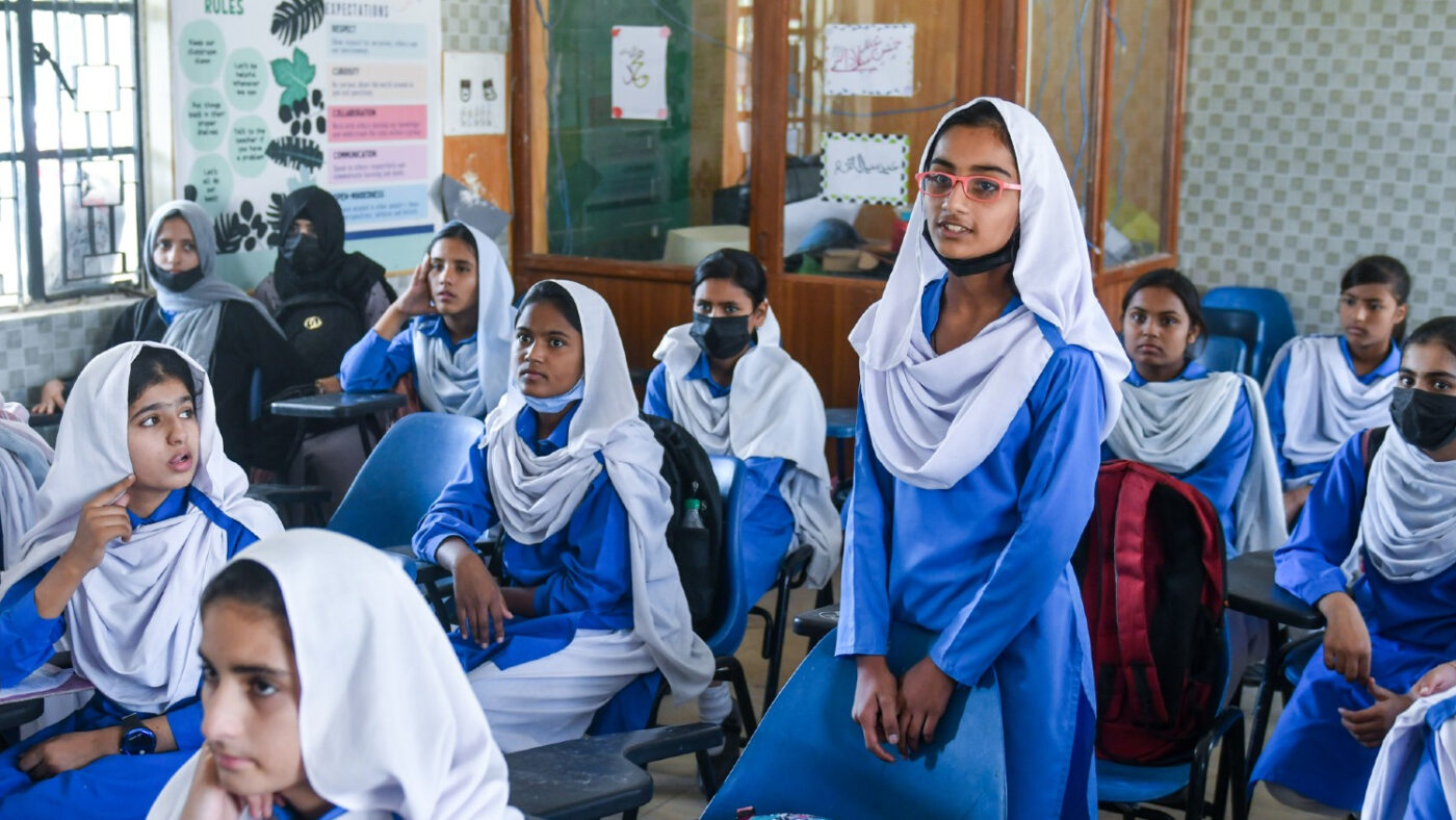 Aakasha and her classmates at school, she is stood up wearing glasses. They are wearing blue long sleeve dresses and white headscarves.