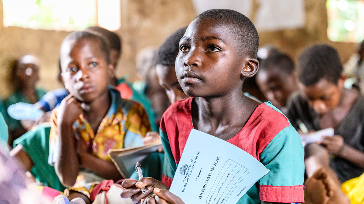 Mary in school with her classmates, holding an exercise book and looking up at the teacher.