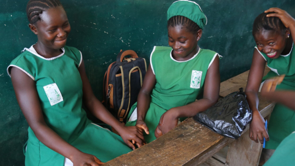 Student Sallay, who has a growth on her left hand, sits with two female friends around a desk. One of the girls is holding Sallay's right hand. They're all wearing school uniform.