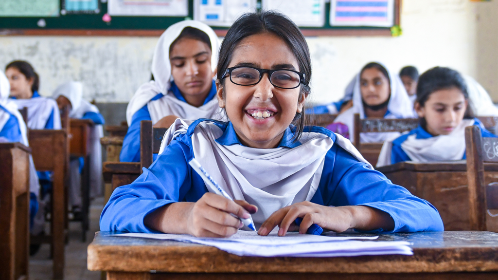 Minahil, who wears spectacles, smiles broadly as she sits at her classroom desk.