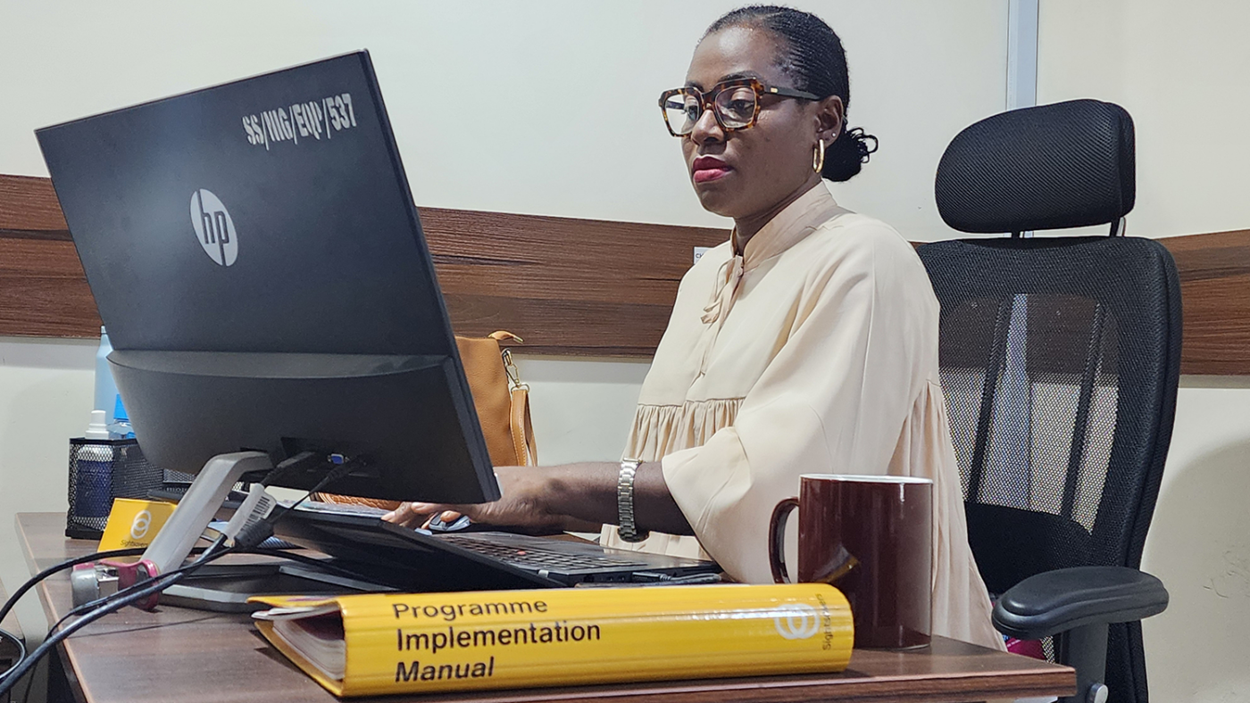 Grace looks at a computer screen while sitting at a desk. A Sightsavers folder is also on the desk.