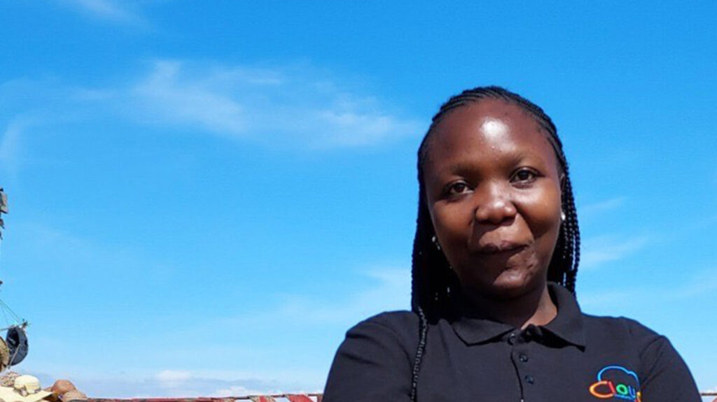 IT Bridge Academy graduate Lyne smiles while folding her arms. Behind her is a bright blue sky.