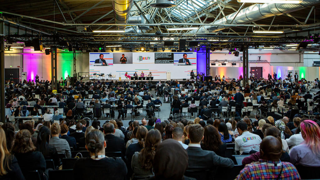 A large audience in a warehouse-like room watches a panel on the Global Disability stage.